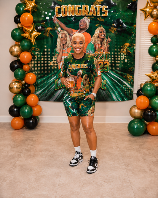 Person in green, orange and gold attire standing in front of a decorated wall with balloons and 'Congrats' sign.