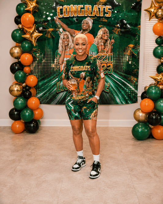 Person in green, orange and gold attire standing in front of a decorated wall with balloons and 'Congrats' sign.